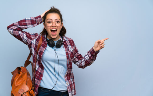Teenager student girl over isolated blue wall surprised and pointing finger to the side Enrol now for the 2025/26 Academic Year!
